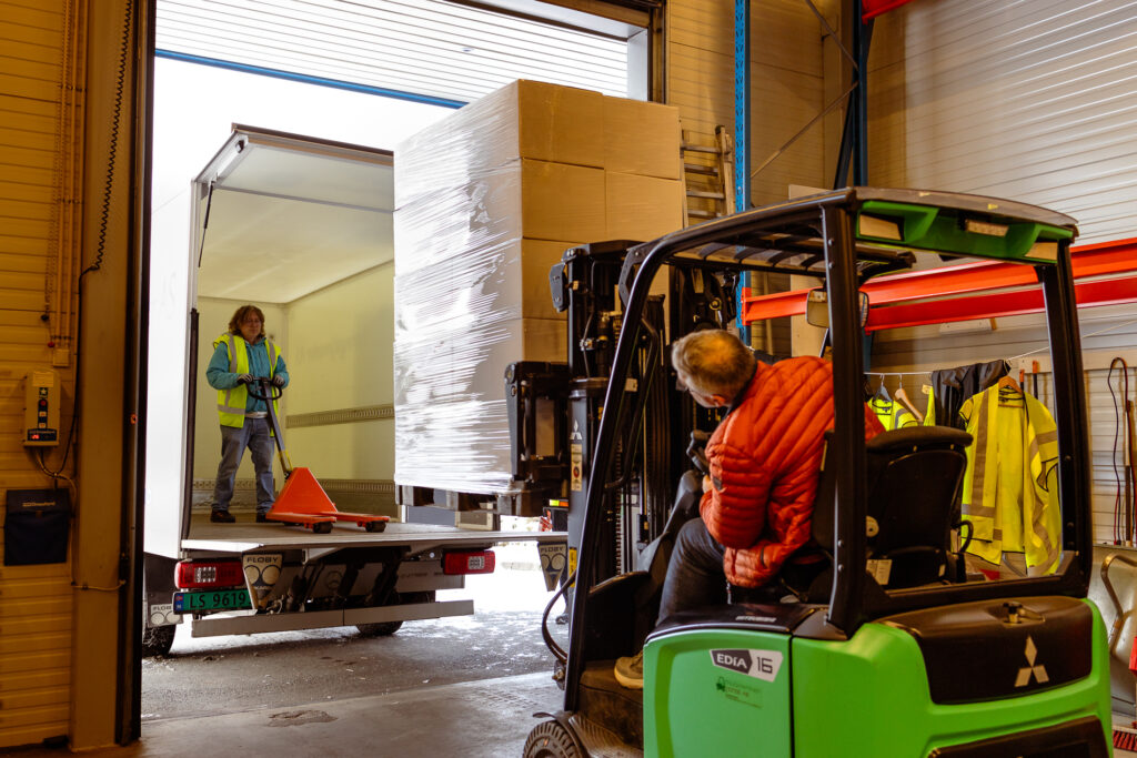 Mannlig medarbeider kjører truck og løfter en pall med esker inn på lastebilen der en annen medarbeider står klar med jekketrall for å ta imot.